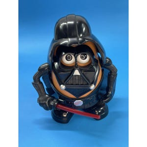 May include: A Darth Vader-themed Mr. Potato Head toy. The toy is primarily black with a black helmet, mask, and cape. It has a tan potato body with white eyes and a red lightsaber. The toy is posed on a blue background.