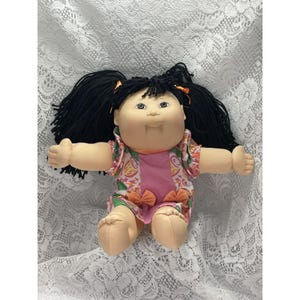 May include: A doll with a round face, tan skin, and black yarn hair styled in two pigtails with orange ribbons. The doll wears a short-sleeved dress featuring a pink center panel and a multi-colored pattern of shapes, accented with two small orange bows at the waist.