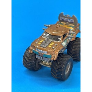 May include: A brown and gold toy monster truck with large black tires. The truck has a decorative blade on the back and the words "Masters of the Universe" on the top. The truck has yellow and green accents and is on a blue background.