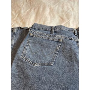 May include: Close-up of blue denim jeans with a classic five-pocket design. The back pocket features a stitched 'W' logo. The jeans are made of a durable denim fabric and have a traditional straight-leg cut.