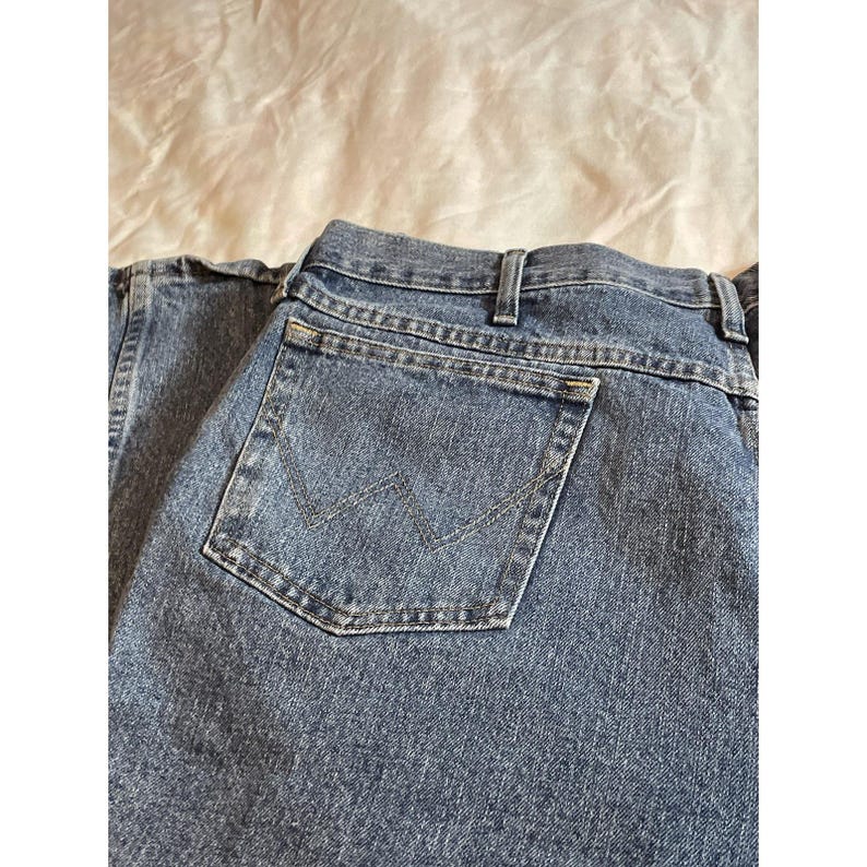 May include: Close-up of blue denim jeans with a classic five-pocket design. The back pocket features a stitched 'W' logo. The jeans are made of a durable denim fabric and have a traditional straight-leg cut.
