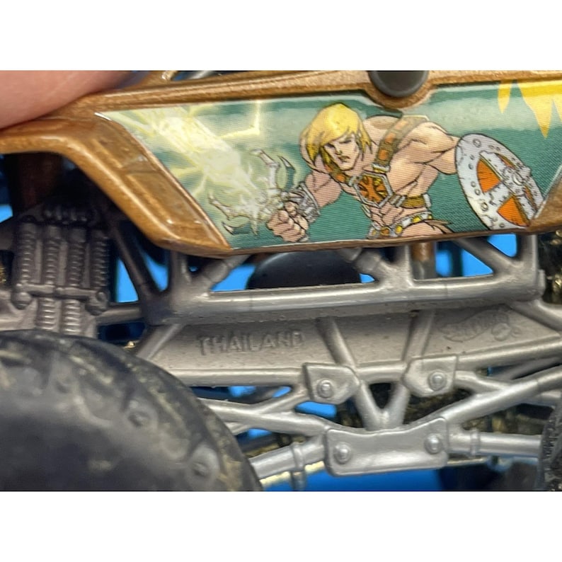 May include: Close-up of a toy monster truck with a gold and green design. The side features a graphic of a muscular blonde man with a shield. The word "THAILAND" is printed on the metal chassis.