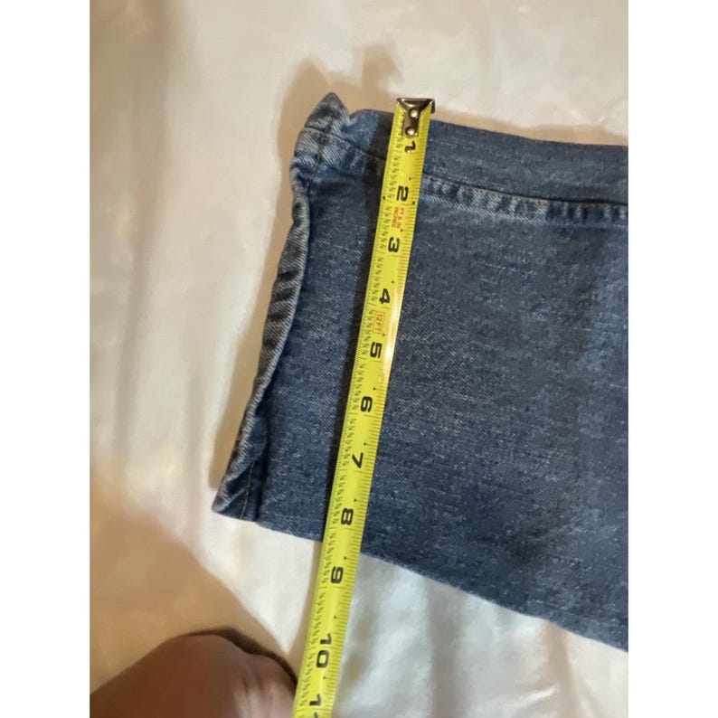 May include: A section of blue denim fabric is measured with a yellow tape measure. The tape measure shows a length of approximately 10 inches. The denim has a classic, faded appearance.