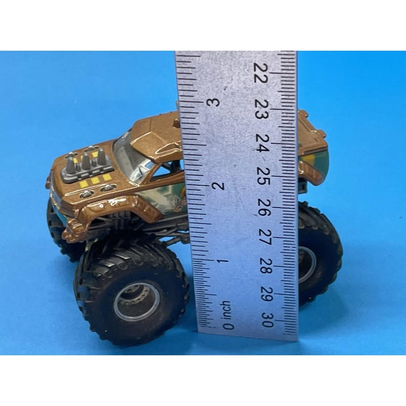 May include: A brown and green monster truck toy with large black tires. The toy is approximately 3 inches (7.6 cm) in height, as measured by a ruler. The truck has a detailed design with a visible engine and cabin.