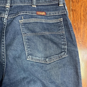 Puede incluir: Primer plano de la parte trasera de unos vaqueros azules. Los vaqueros tienen un bolsillo trasero y la etiqueta roja de la marca "Wrangler". El denim tiene un aspecto cl&aacute;sico y desgastado.