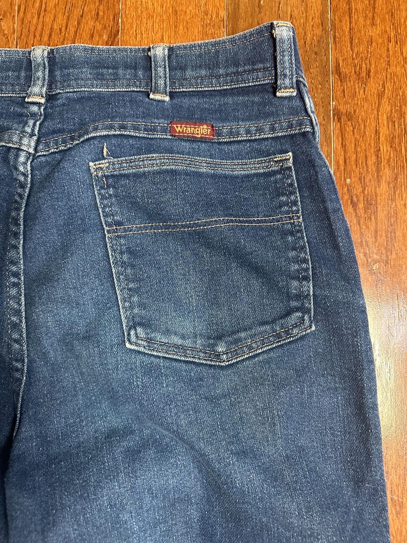Puede incluir: Primer plano de la parte trasera de unos vaqueros azules. Los vaqueros tienen un bolsillo trasero y la etiqueta roja de la marca "Wrangler". El denim tiene un aspecto cl&aacute;sico y desgastado.