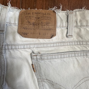 May include: Close-up of light-wash denim jeans with a frayed hem. A brown leather patch with the Levi Strauss & Co. logo and text is visible. The jeans have a back pocket and a button closure.