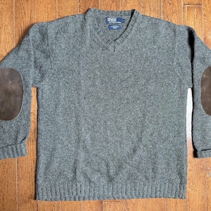 May include: A gray V-neck sweater with brown suede elbow patches. The sweater has a ribbed hem and cuffs. The Polo Ralph Lauren label is visible at the neckline. The sweater is made of a thick knit material.