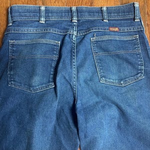 Puede incluir: Primer plano de la parte trasera de unos vaqueros azules. Los vaqueros tienen dos bolsillos traseros, trabillas y una peque&ntilde;a etiqueta roja de Wrangler. La mezclilla tiene un aspecto cl&aacute;sico y desgastado.