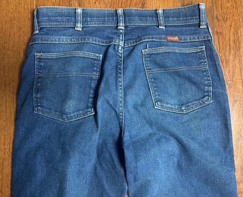Puede incluir: Primer plano de la parte trasera de unos vaqueros azules. Los vaqueros tienen dos bolsillos traseros, trabillas y una peque&ntilde;a etiqueta roja de Wrangler. La mezclilla tiene un aspecto cl&aacute;sico y desgastado.