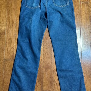 Puede incluir: Un par de vaqueros de mezclilla azul, vistos desde la parte trasera, con dos bolsillos traseros y una cinturilla. Los vaqueros est&aacute;n colocados sobre una superficie de madera, mostrando su dise&ntilde;o cl&aacute;sico y detalles de costura.
