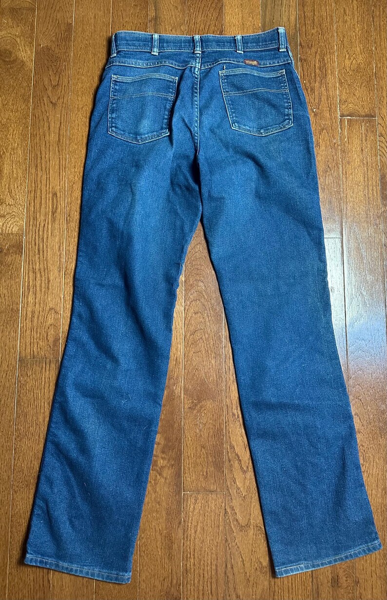 Puede incluir: Un par de vaqueros de mezclilla azul, vistos desde la parte trasera, con dos bolsillos traseros y una cinturilla. Los vaqueros est&aacute;n colocados sobre una superficie de madera, mostrando su dise&ntilde;o cl&aacute;sico y detalles de costura.