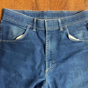 Puede incluir: Primer plano de unos vaqueros de mezclilla azules. Los vaqueros tienen un dise&ntilde;o cl&aacute;sico de cinco bolsillos con cierre de bot&oacute;n y cremallera. La mezclilla tiene un lavado descolorido y los bolsillos tienen un forro blanco. Los vaqueros son de talle medio.