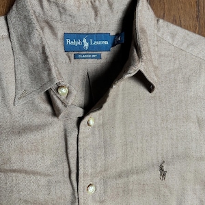 May include: A tan button-down shirt with a herringbone pattern. The shirt features a classic collar, buttoned cuffs, and a small embroidered logo of a polo player. The Ralph Lauren label is visible.