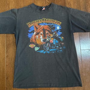 May include: Dark gray Harley-Davidson t-shirt featuring a wolf and a motorcycle graphic. The design includes the Harley-Davidson logo and a rider on a red motorcycle under a full moon. The shirt is short-sleeved.