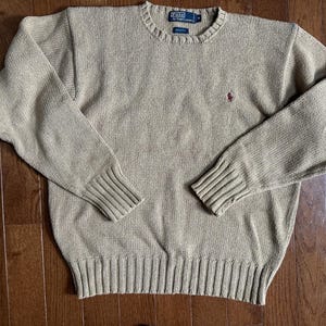 May include: A tan crew neck sweater with long sleeves. The sweater has a ribbed hem and cuffs, and a small red logo on the chest. The brand label is visible at the neckline. The sweater is made of a knit fabric.