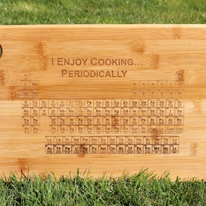 Op de afbeelding: Een rechthoekige bamboe snijplank met een gat in de linkerbovenhoek. De plank is gegraveerd met de tekst "I ENJOY COOKING... PERIODICALLY" boven een periodiek systeem der elementen.