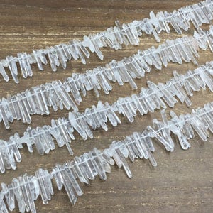 May include: A strand of clear quartz crystal points, arranged in a staggered pattern, on a wooden surface. The crystals are various lengths and have a natural, raw finish.