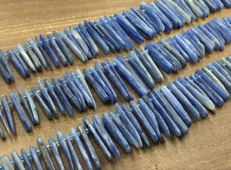 May include: A strand of blue kyanite beads, each bead is a long, thin, and slightly irregular shape. The beads are strung on a thin wire and are arranged in a staggered pattern.