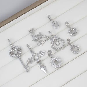 May include: A collection of silver-toned pendant charms in various designs, including a key, cross, and floral shapes. Some charms feature clear stone settings, while others have open frames for customization. The pendants are displayed on a white surface.