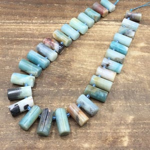 Pu&ograve; includere: Una collana di perline di amazonite di colore blu chiaro, verde e marrone infilate su un cordoncino blu. Le perline sono cilindriche e hanno una finitura liscia e lucida.