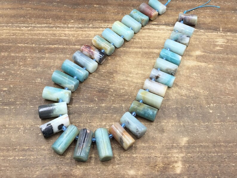 Pu&ograve; includere: Una collana di perline di amazonite di colore blu chiaro, verde e marrone infilate su un cordoncino blu. Le perline sono cilindriche e hanno una finitura liscia e lucida.