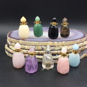 Pode incluir: Dez pequenas garrafas de pedras preciosas com tampas douradas. As garrafas são de várias cores, incluindo rosa, verde, branco, preto e azul. As garrafas estão dispostas em círculo sobre uma fatia de ágata branca e marrom.