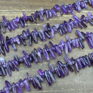 May include: A strand of amethyst gemstone beads, each bead is a long, thin, pointed shape, resembling a spike or a teardrop. The beads are a deep purple colour and are strung together on a clear thread.
