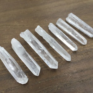 May include: Eight clear quartz crystal points, also known as wands, are arranged in a row on a wooden surface. The points are all approximately the same size and shape, and they have a natural, unpolished finish.