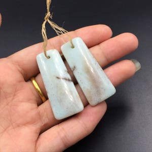 Puede incluir: Dos colgantes rectangulares de piedra preciosa azul claro con vetas marrones, suspendidos por una cuerda de fibra natural. Los colgantes se sostienen en una mano, con un anillo de oro visible. El fondo es de color oscuro.