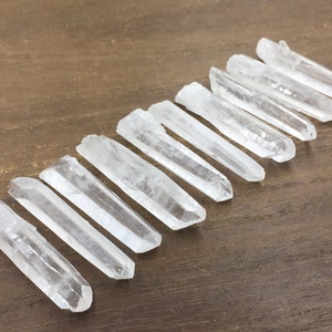 May include: A collection of ten clear quartz crystal points, arranged in a row on a brown wooden surface. The points are all roughly the same size and shape, and they have a natural, unpolished finish.