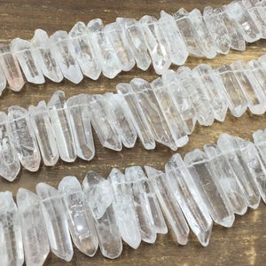 May include: Strands of clear quartz crystal points, strung together. The crystals are irregularly shaped with a natural, raw appearance. The quartz has a translucent quality, with some areas appearing more opaque. The crystals are strung on a thin, clear cord.