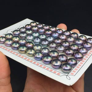 Puede incluir: Primer plano de una tarjeta blanca con un patrón de cuadrícula, que muestra una colección de perlas negras iridiscentes. Las perlas están dispuestas en filas y columnas, mostrando sus variaciones de color únicas.