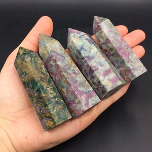 Może przedstawiać: Cztery zielone, brązowe i fioletowe szpiczaste kamienie szlachetne, znane również jako różdżki lub wieże, są ułożone w rzędzie na dłoni. Szpiczaste kamienie są wypolerowane i mają naturalny, organiczny wygląd.