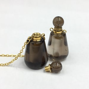 Può includere: Due flaconi di profumo in vetro marrone con tappi dorati e una catena dorata. Un flacone è su una catena e l'altro è fuori dalla catena. Il flacone sulla catena ha un tappo dorato con una piccola apertura. Il flacone fuori dalla catena ha un tappo rotondo dorato.