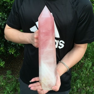 Op de afbeelding: Een grote, roze en witte kristallen punt, ook wel een kristallen staf genoemd, wordt vastgehouden door de handen van een persoon. De kristal is ongeveer 30 cm lang.