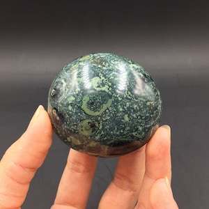 May include: A green and black sphere with a swirling pattern of white and green spots. The sphere is being held in a hand.
