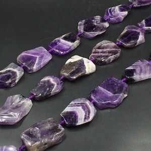Puede incluir: Una hebra de cuentas de piedra preciosa de amatista púrpura, cada cuenta tiene una forma irregular única con una textura natural y rugosa. Las cuentas están enhebradas en un cordón púrpura.