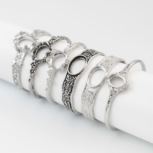 Op de afbeelding: Een verzameling zilverkleurige metalen armbanden met diverse decoratieve ontwerpen. De armbanden hebben open instellingen voor stenen of edelstenen, met ingewikkelde patronen en texturen. De ontwerpen omvatten bloemen-, textuur- en opengewerkte stijlen, met een reeks versieringen.