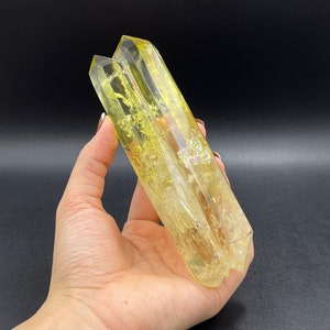 Op de afbeelding: Een dubbel geëindigd helder kristal met gele en gouden insluitsels. Het kristal wordt in een hand gehouden.