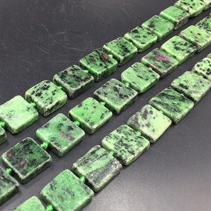 May include: A strand of green gemstone beads with black and brown flecks. The beads are square shaped and strung on green thread.