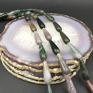 Può includere: Una collana di perle di agata verde, marrone e viola a forma di lacrima. Le perle sono disposte su una fetta di agata.