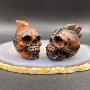 Peut inclure: Deux sculptures de crânes en pierre rouge et noire sur une tranche d'agate blanche. Les crânes sont détaillés et présentent un motif en pointe sur le dessus.