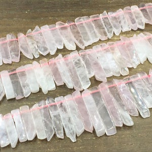 May include: A strand of pink quartz crystal beads, each bead is a long, thin, and slightly irregular shape. The beads are strung on a pink thread.
