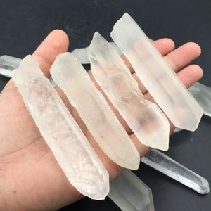 May include: Several clear quartz crystal points of varying sizes are displayed. The crystals are held in a hand against a dark background. The quartz crystals have a natural, rough texture and are translucent.