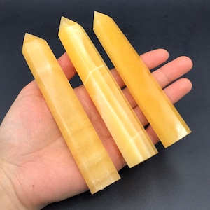 Könnte beinhalten: Drei goldgelbe Calcit-Kristallstäbe mit spitzen Spitzen. Die Stäbe werden in einer Hand vor einem dunklen Hintergrund gehalten. Die Kristalle haben ein glattes, poliertes Aussehen und sind rechteckig.