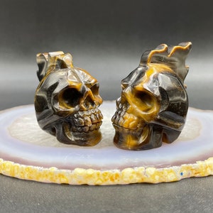 May include: Two tiger's eye skull sculptures, one with a mohawk, on a white and gold agate slice. The skulls are carved from brown and gold tiger's eye stone.