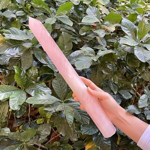 Op de afbeelding: Een grote, roze, rozenkwarts kristallen punt, ook wel een toverstaf genoemd, wordt in een hand gehouden. Het kristal is glad en gepolijst, met een puntige punt. De achtergrond is een groene, bladerrijke plant.