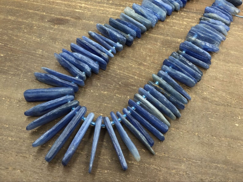 May include: A strand of blue kyanite beads, each bead is a long, thin, and slightly irregular shape. The beads are strung together with a thin, clear thread.