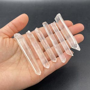 May include: A collection of seven clear quartz crystal points, also known as wands, displayed in a hand. The points are various lengths and thicknesses.